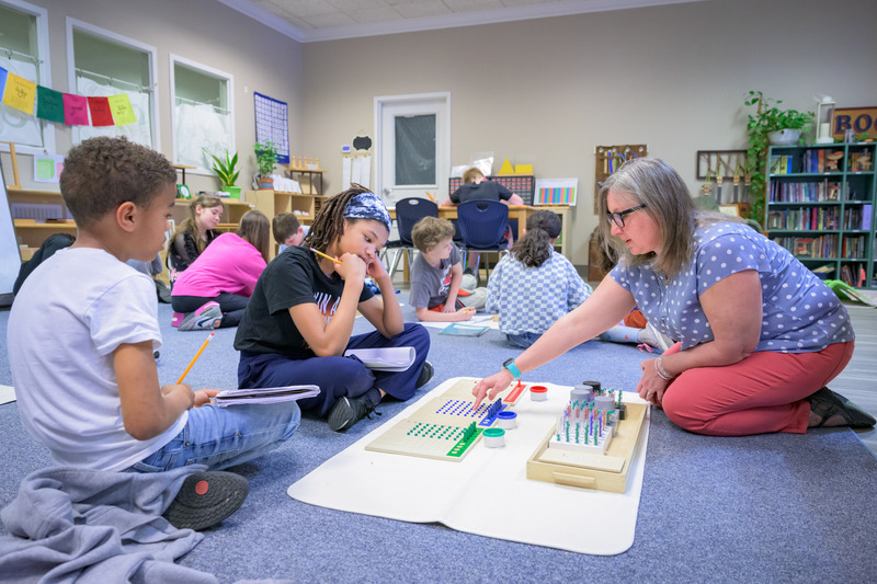 A teacher sits on the floor with her young students, talking them through a project together at First State Academy Montessori school.