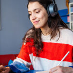 A woman wearing a red and white striped sweater and headphones, is sitting at a desk with her laptop, writing in a notebook with a pencil. Behind the individual is a bookshelf filled with various books, and above it hangs colorful artwork featuring a face. The setting suggests a cozy study environment.