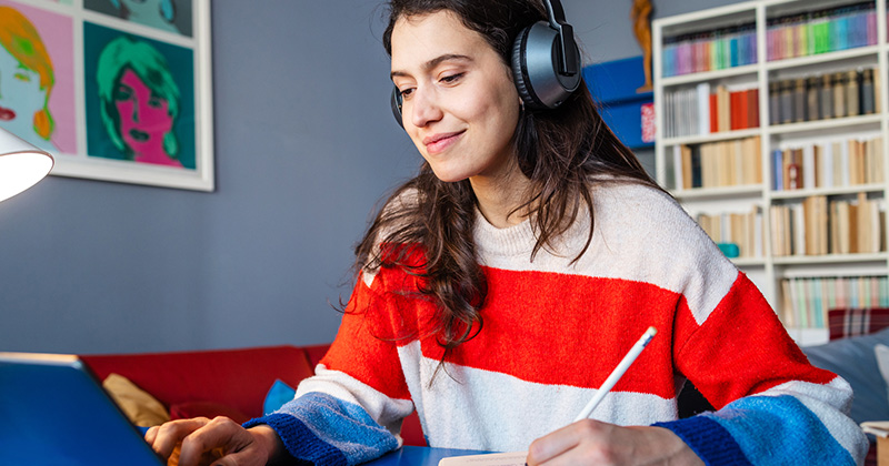 A woman wearing a red and white striped sweater and headphones, is sitting at a desk with her laptop, writing in a notebook with a pencil. Behind the individual is a bookshelf filled with various books, and above it hangs colorful artwork featuring a face. The setting suggests a cozy study environment.