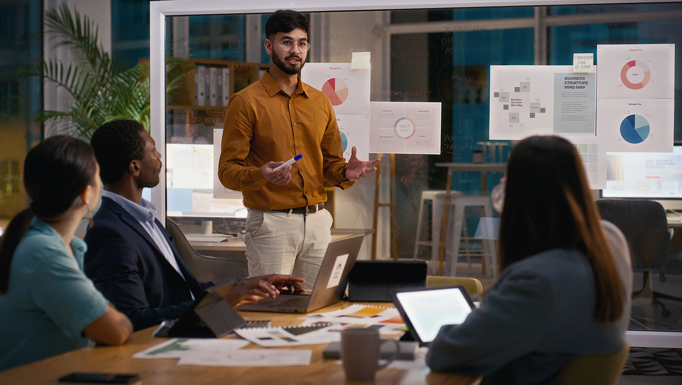 Standing man giving presentation at meeting to people sitting around a table