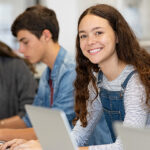 Happy young high school student working at her laptop in a classroom, looking at the camera.