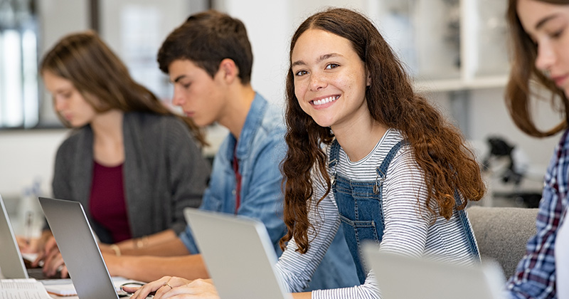 Happy young high school student working at her laptop in a classroom, looking at the camera.