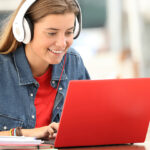 Young girl sitting at a kitchen table working on her laptop with headphones on.